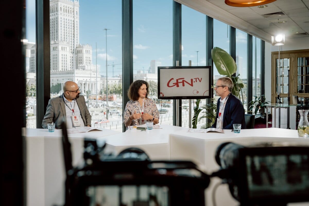 Three people sit at a panel discussion in a studio with large windows overlooking the city skyline. Behind them a screen with the words "GFN.TV" is projected. The man on the left and the man on the right listen intently to the woman in the middle speak, while a photographer at the conference captures the moment.  