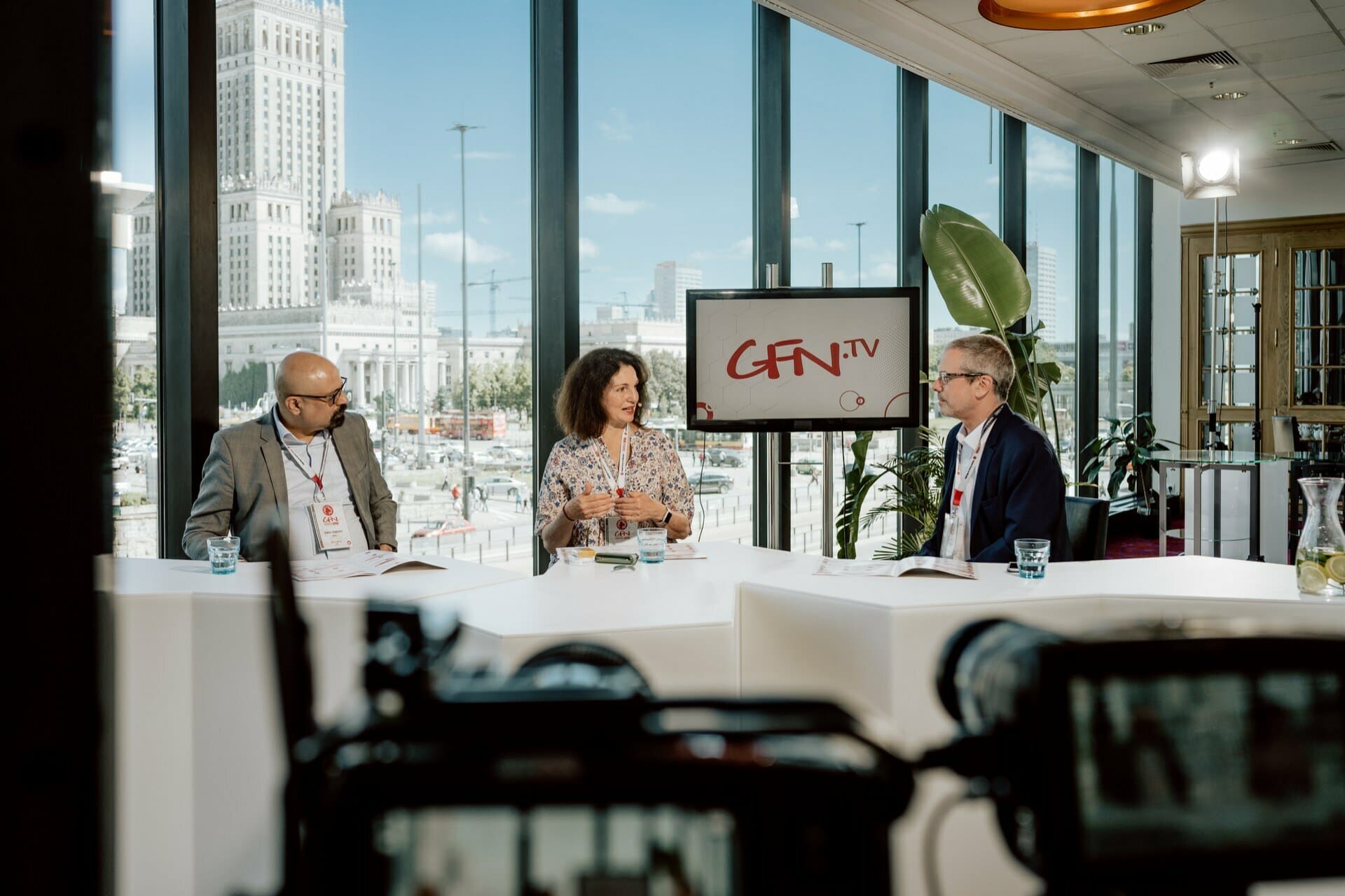 Trzy osoby siedzą przy panelu dyskusyjnym w studiu z dużymi oknami wychodzącymi na panoramę miasta. Za nimi wyświetlany jest ekran z napisem „GFN.TV”. Mężczyzna po lewej i mężczyzna po prawej słuchają uważnie wypowiedzi kobiety pośrodku, a fotograf na konferencje uwiecznia ten moment.