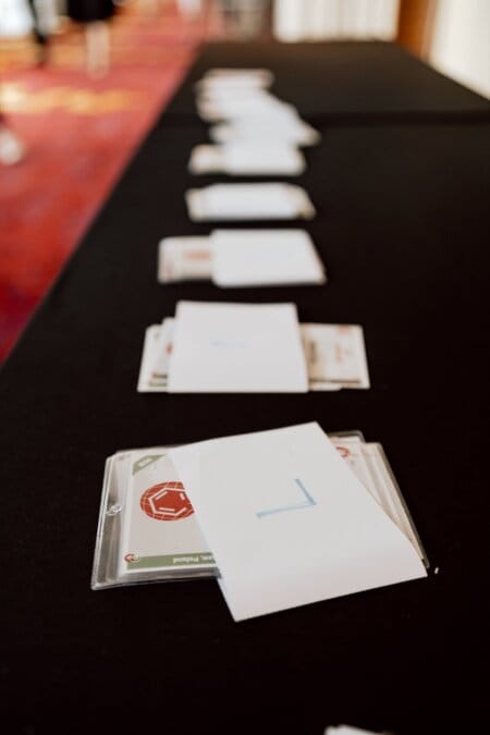 A row of white envelopes placed on a black tablecloth. Each envelope appears to contain a card or document that is only partially visible, and some are marked with blue numbers. In the background you can see a smudge of red carpet and bright scenery, reminiscent of scenes captured by a photographer at conferences.  