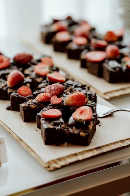 A tray of chocolate brownies sprinkled with fresh strawberries and topped with chocolate sauce. A spatula is placed under one of the brownie pieces. The tray and its goodies rest on parchment paper and are ready to be captured by the photographer at the conferences in the background.  