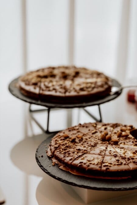 Two chocolate pizzas are displayed on black slate cake stands. Each pizza is covered with chocolate glaze and sprinkled with breadcrumbs. One pizza is partially sliced and missing a piece, while the other is intact. The arrangement looks so elegant that it could be captured by a photographer at conferences. Both are placed on a white surface.    