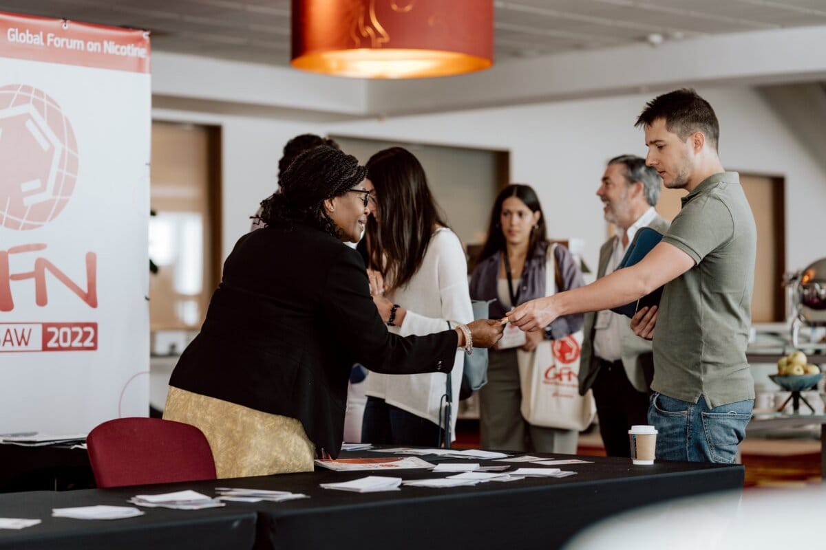 A woman in a black jacket and glasses hands a badge to a man in a green shirt at the registration booth, with several other people waiting in line behind them. A banner with the words "Global Forum on Nicotine" is visible in the background, capturing the perfect moment for any photographer at the conference. 