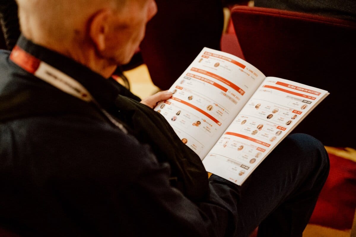 A person is sitting, holding and reading an open brochure with the event program. The brochure consists of several pages containing a mix of text and images, probably including a detailed schedule of the conference and speakers. The person wears a lanyard with a badge around his neck, perhaps preparing for a photo essay of the conference.  