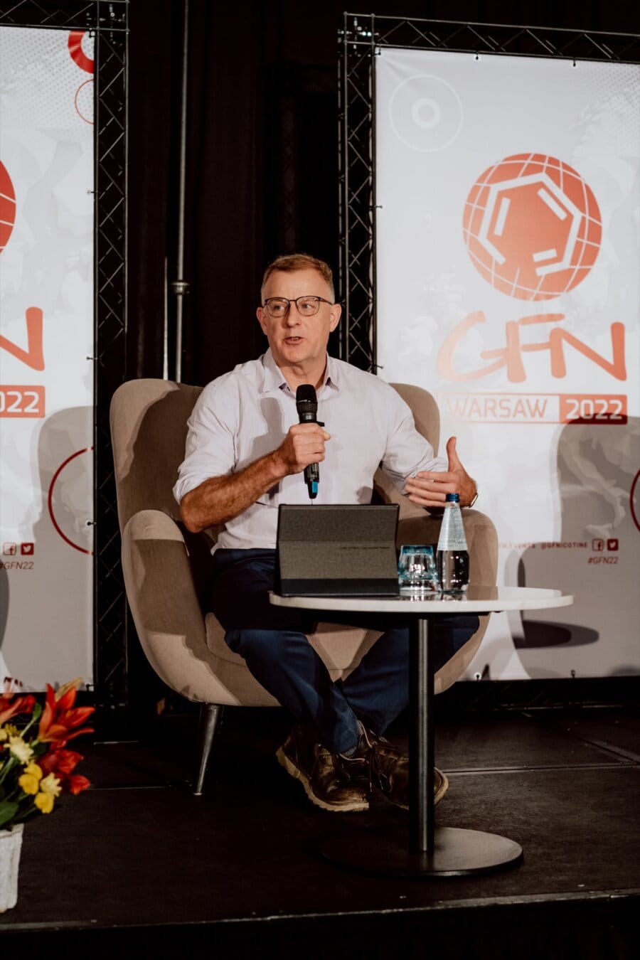 A man in a white shirt and dark pants speaks into a microphone while sitting in a chair on stage. He has a laptop on his lap, and in front of him is a small table with a bottle of water. Behind them are banners with the words "CTN Warsaw 2022," perfectly capturing the moment of the conference photo-op.  