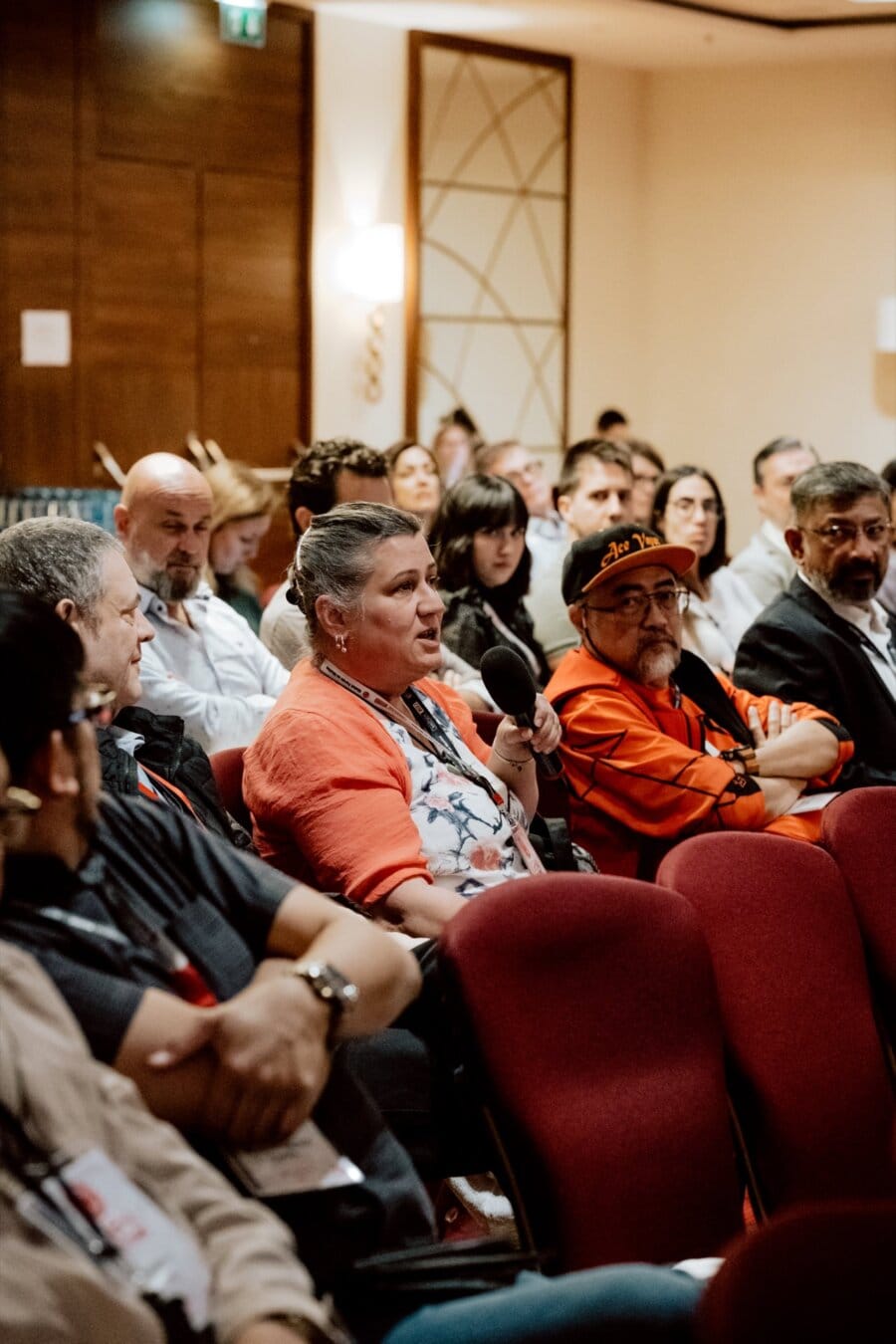 At a conference or seminar, a group of people sit in rows. The person in the middle of the photo, wearing an orange sweater, holds a microphone and gives the impression of speaking or asking a question. This moment was perfectly captured by Marcin Krokowski, an acclaimed event photographer. Others around them focus on the speaker, listening intently.   