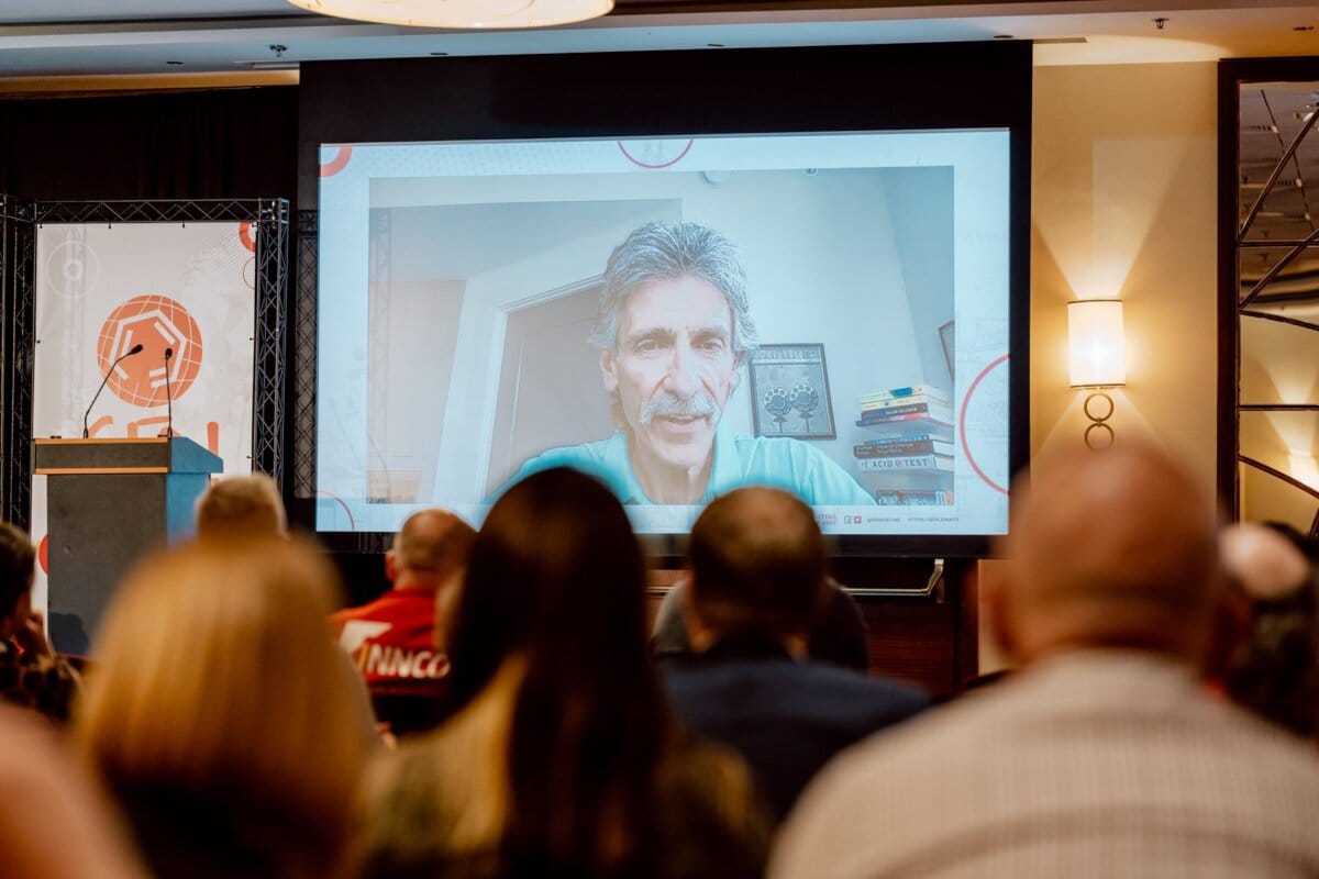 A group of people are sitting in the audience facing a large screen at the front of the conference room. The screen shows a man with gray hair and a mustache speaking, while books and artwork are visible in the background. To the left of the screen is a podium, creating an engaging photo essay of the conference.  