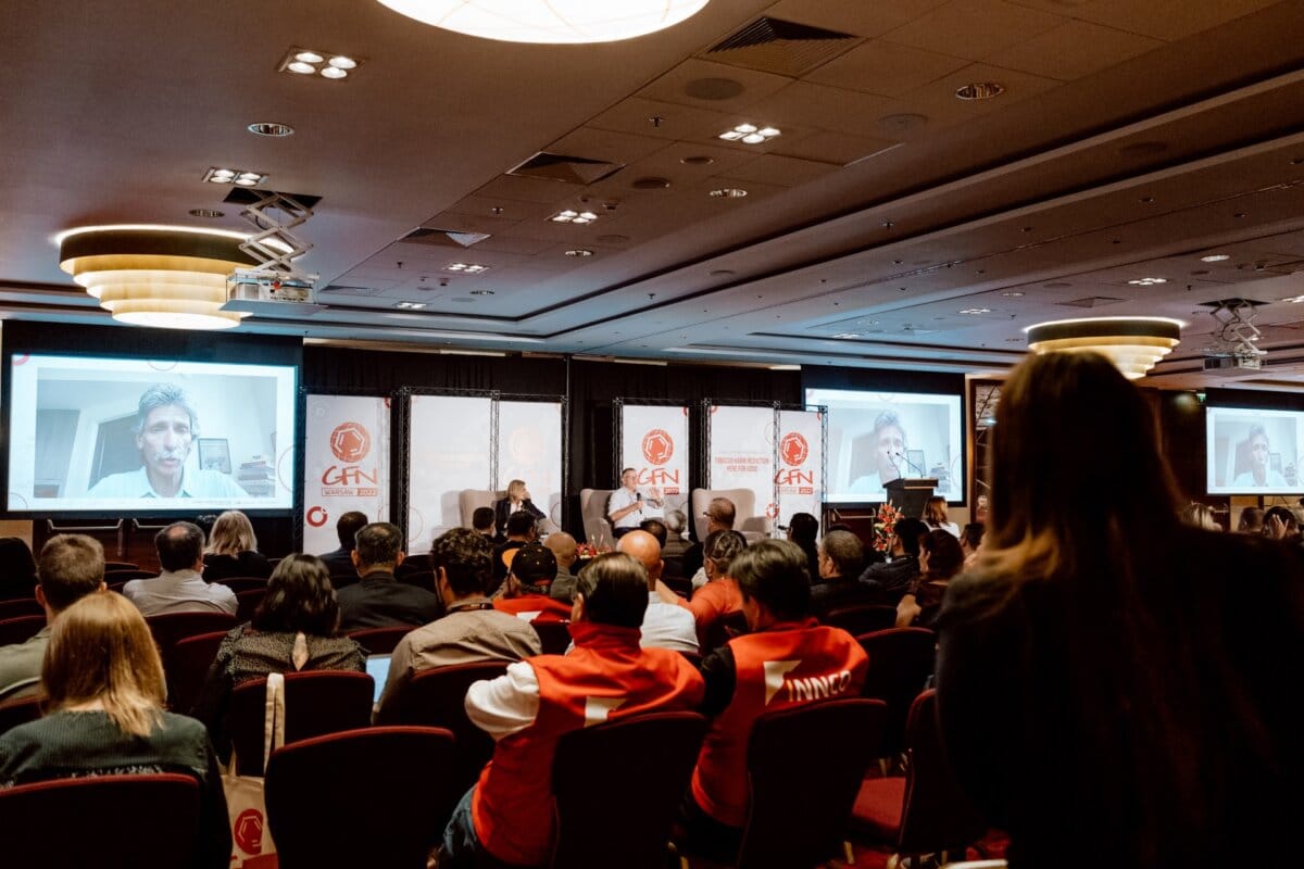 A conference room filled with attendees watching video streamed live on three large screens. The screens show a speaker, and behind a panel of two speakers are banners with logos. A photo report from the conference shows seated spectators, some wearing red T-shirts.  