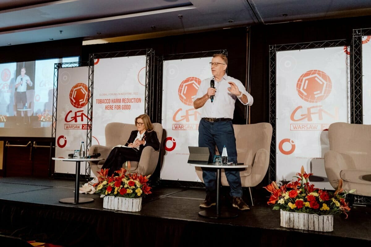 A man stands and speaks into a microphone on stage during a conference, while a woman sits next to him and listens intently. Behind them hang banners reading "Tobacco harm reduction: here for good" and "GFN, Warsaw." On the screen to the left is a presentation showing a photo essay of the conference.  