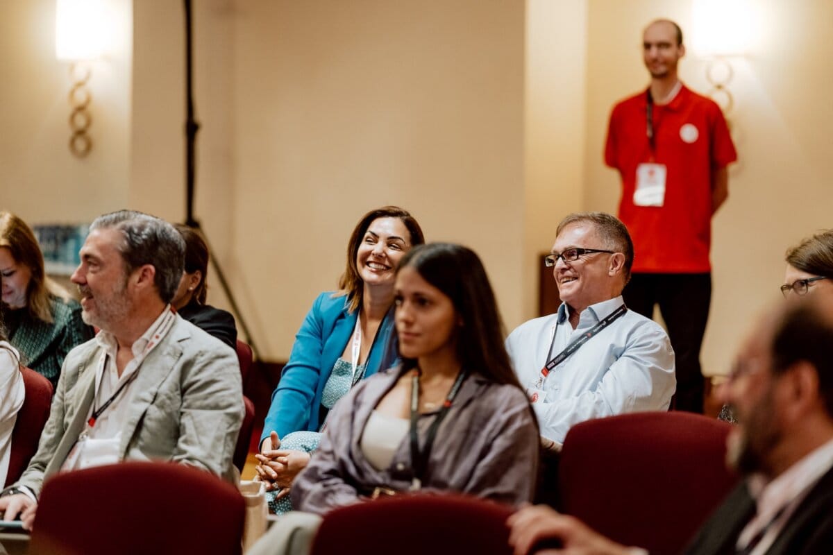 A group of people dressed in both business and casual attire are sitting in a seminar room. They are smiling and give the impression of being involved in the event, which is a perfect fit for a photo essay of the conference. In the background stands a man in a red shirt and lanyard.  