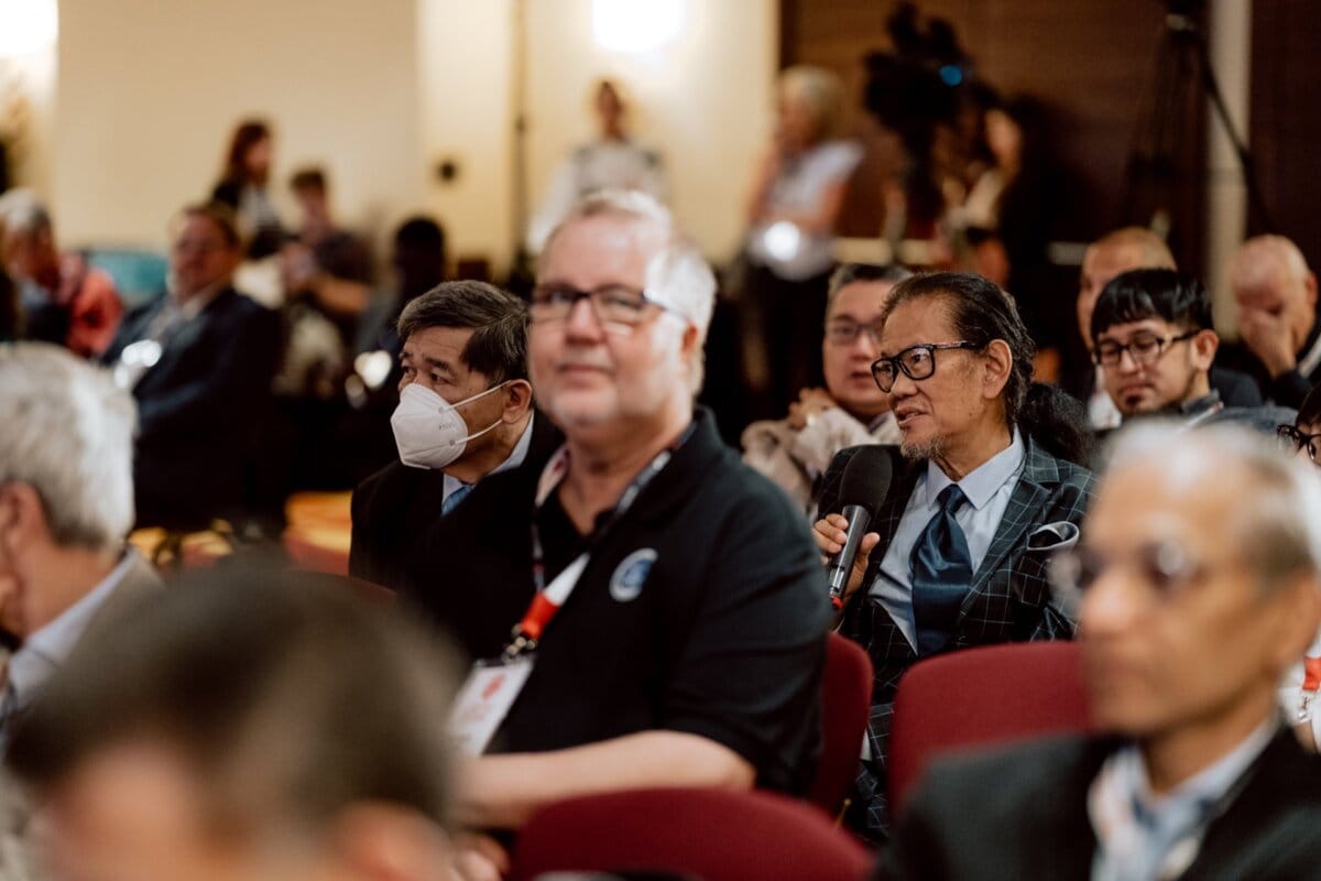 A man dressed in a suit and holding a microphone is speaking at the event. He is surrounded by other attendees sitting in rows, some wearing masks. More people can be seen in the background, some standing, others sitting, with a camera set up to record this photo essay of the conference.  