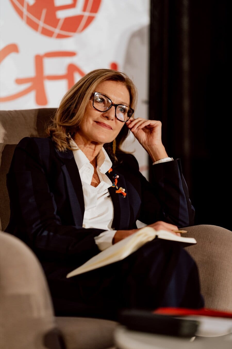 A person wearing glasses, dressed in a dark jacket and white shirt, sits on a chair and holds a notebook. They have a thoughtful expression on their face and touch their temples with one hand. The scene appears as part of a photo essay of the conference, with a fuzzy banner in the background.  