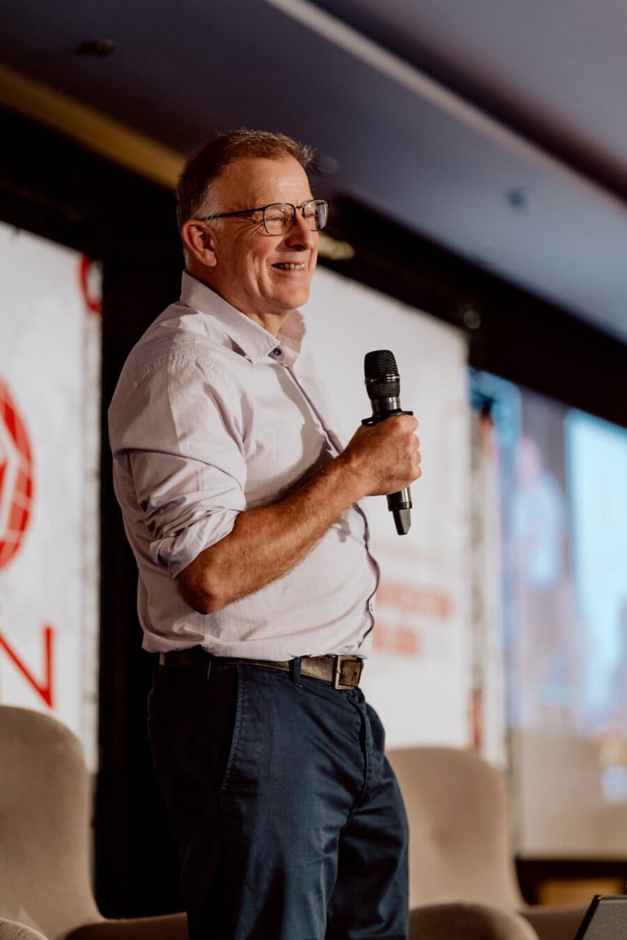 A man wearing glasses, a white shirt and dark pants is standing on stage, smiling and holding a microphone. He appears to be giving a speech or presentation. In the background is a banner with partially visible text that captures the essence of the event in this vibrant photo essay of the event.  