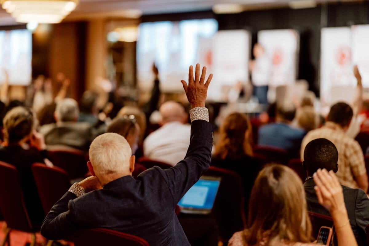 A conference room filled with participants sitting facing the speaker on stage. Several people raise their hands, indicating participation. The room has a professional setting with a large screen displaying visual content, ideal for photo coverage of the conference to capture the immersive atmosphere.  