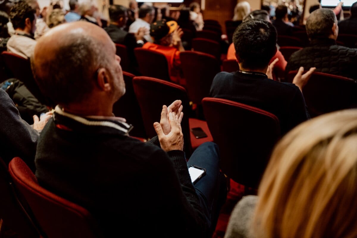 Spectators sitting in rows of red chairs clap, engaged in what appears to be a formal event or presentation. The focus is on the backs of those in the foreground. Some of the participants have phones or other small objects in their laps, captured as part of a bustling reportage of the event.  