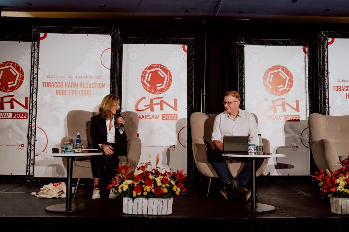 Two people sit on stage during a panel discussion at the World Nicotine Forum 2022 in Warsaw, Poland. In the background are event banners with the forum logo and theme: "Tobacco harm reduction: here for good." Red flowers adorn the stage, adding to the reportage of the event.  