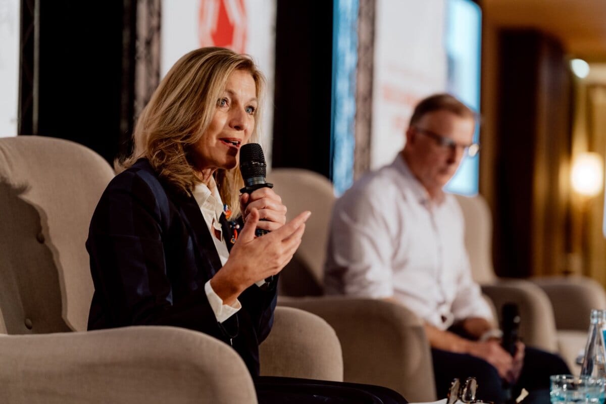 A woman holds a microphone and speaks while sitting in a chair on stage during a panel discussion. A man sits next to her and listens attentively. Both are dressed in business attire. In the background are event banners and a blurry audience, perfectly captured in this photo essay of the conference.   