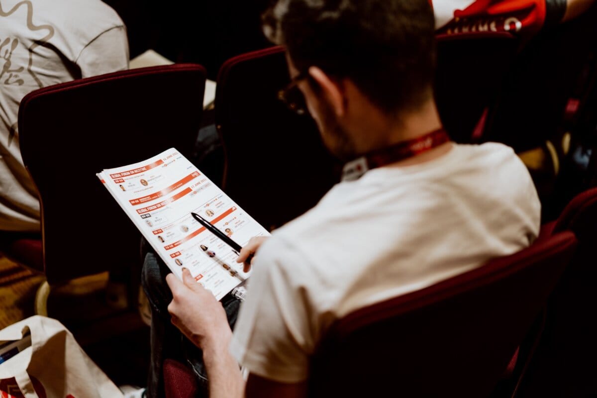 In a chair sits a person wearing glasses and a white shirt, absorbed in reading a program or brochure. The scenery suggests that this is a photo essay of a conference; in the background you can see other participants sitting in rows. 