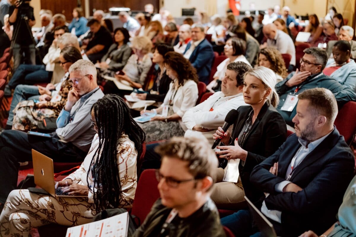 A group of people sitting in rows at a conference or seminar. Many are working at laptops or taking notes. The woman in the foreground is holding a microphone and appears to be asking a question. The atmosphere is attentive and focused, which is perfectly captured by this photo report from the conference.   