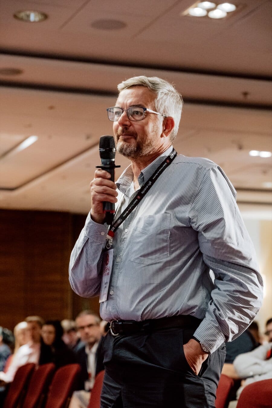 A man with short gray hair and glasses speaks into a microphone during a conference. He is wearing a striped shirt with a visible lanyard and keeps one hand in his pocket. The audience captured in the photo essay of the conference sits in red chairs in the background.  