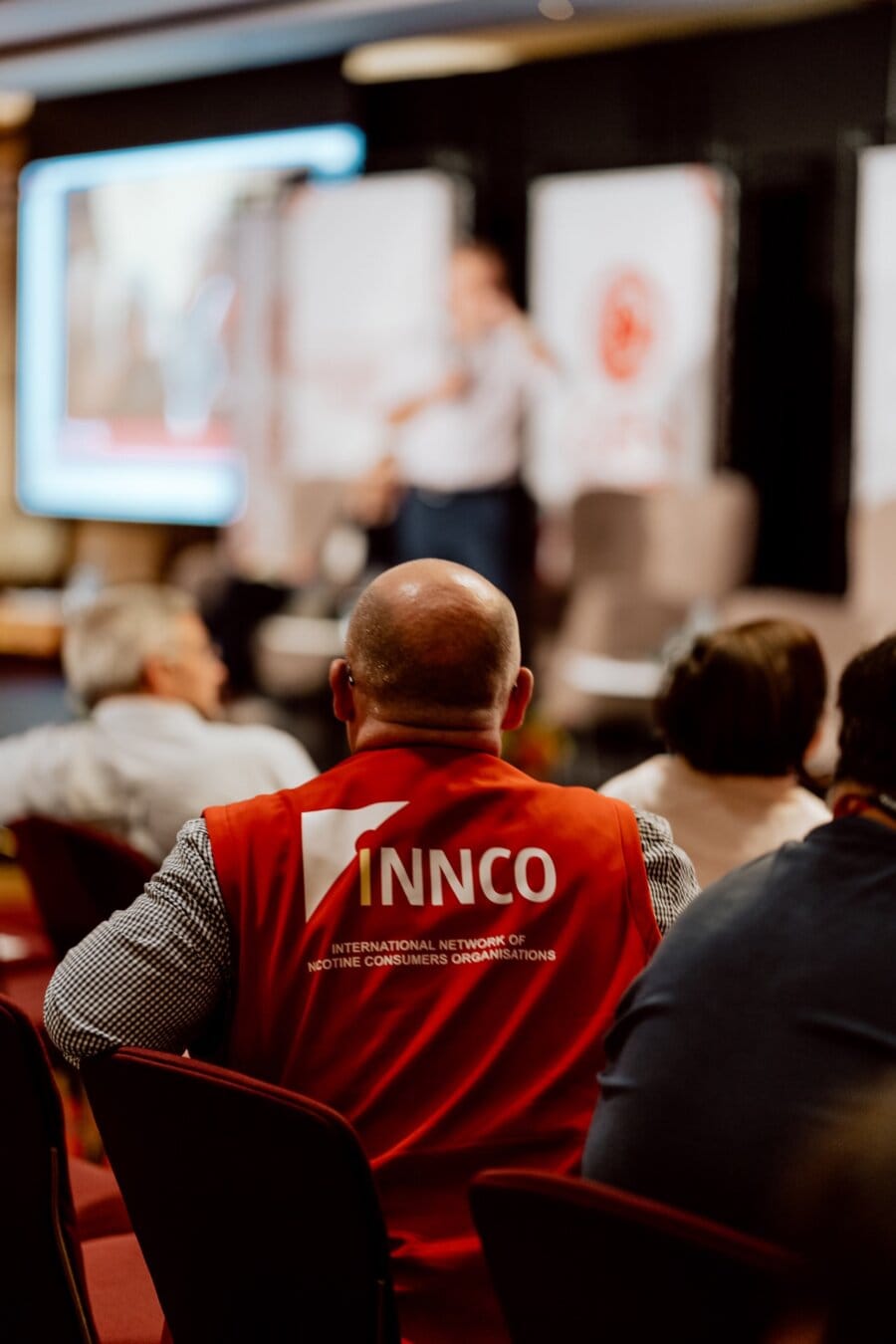 A man sitting in the audience is wearing a red vest with "INNCO" and "International Network of Nicotine Consuming Organizations" printed on the back. In the background are blurred figures and presentation screens, capturing a photo-reportage of an event or conference. 