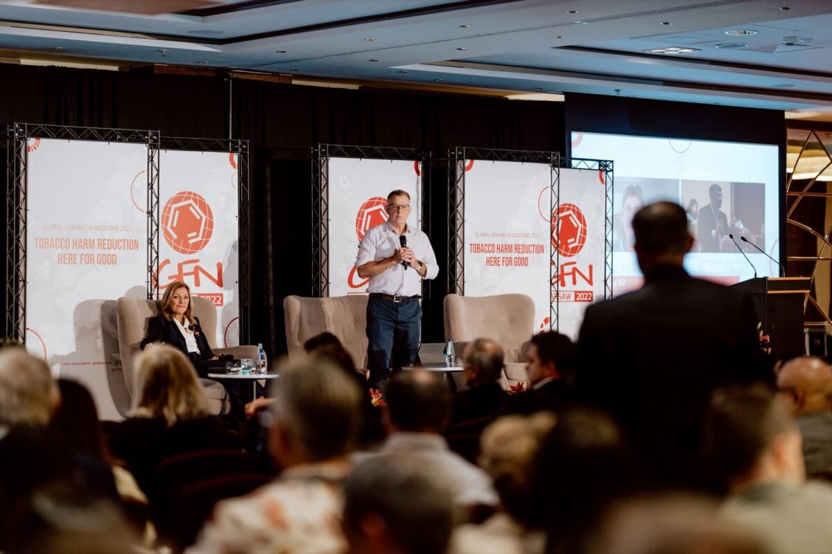 A man stands on stage with a microphone and speaks to the audience. To his right sits a woman. A large screen and posters display the text "Good tobacco harm reduction." Several people can be seen in the audience, one of whom is standing facing the stage and capturing a photo essay of the conference.   