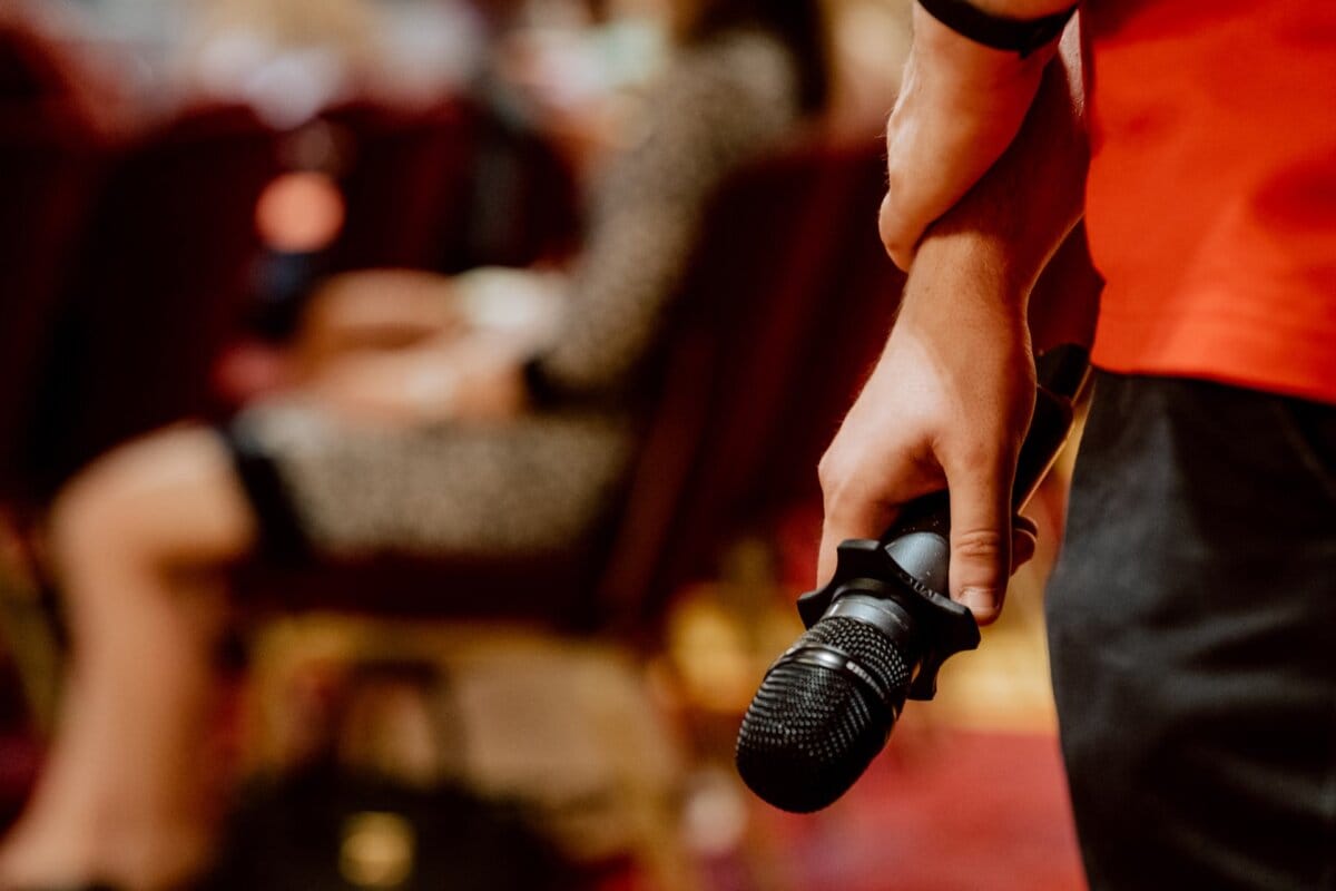 The person holds the microphone close to his hip, standing in a room with rows of fuzzy seated participants in the background, capturing the essence of the photo report of the event. The person is wearing a red shirt, and his hand lightly squeezes the microphone pointing downward. 