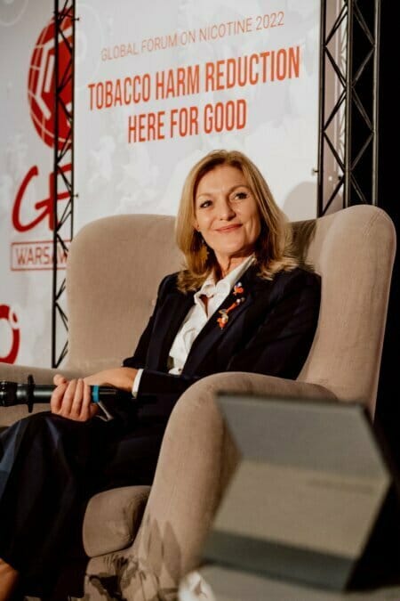 A woman in a dark suit sits in a chair, holding a microphone and smiling. Behind her is a banner with the words "Global Forum on Nicotine 2022" and "Reducing the harms of tobacco: here for good." This photo is part of a photo essay from the conference documenting important discussions at the event.  