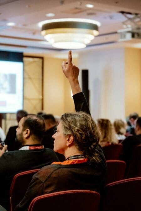 A person raises his hand in a conference room filled with engaged participants. A modern chandelier illuminates the room, while a large screen displays a presentation at the front. This captivating moment is part of a photo essay of the event that captures the essence of the dynamic event.  