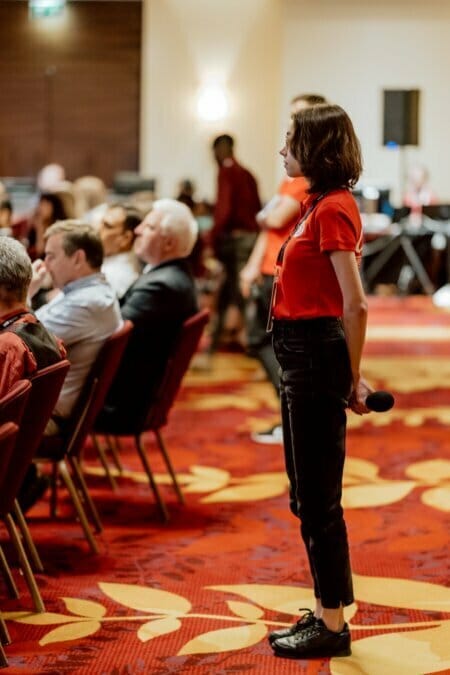 A woman in a red shirt and black jeans stands on the right, holding a microphone and looking toward the seated audience in the conference room. The audience sits in rows of red chairs on a red carpet with yellow leaf patterns, perfect for a detailed photo essay of the conference. 