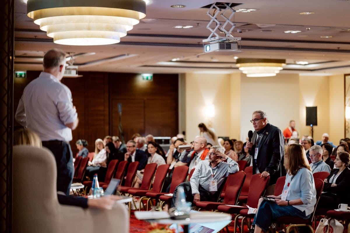 A man stands holding a microphone and speaks to an audience seated in a conference room. Another person faces him at the front. The room has red chairs, large ceiling lights and a projector. Participants listen attentively as a reportage of the event is recorded by the event photographer.   