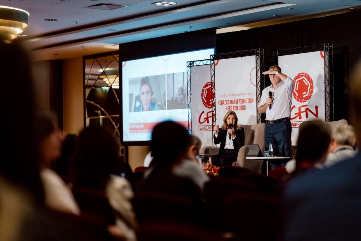 Conference with a man and woman speaking on stage. A sign in the background reads "GFN." A man wearing a hat stands and a woman sits next to him. Behind them is a large screen showing another person participating in a video call. The audience is intently watching the photo-recording of the conference.    