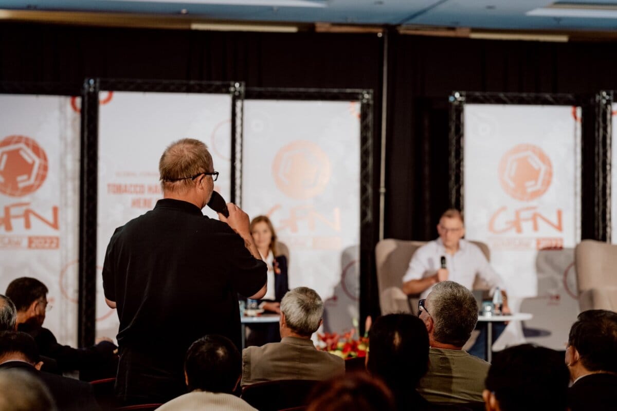 At a conference, a person stands up and asks a question into a microphone. Two panelists sit on the stage in the background, facing the audience. Attendees sit and listen attentively, capturing every detail for an in-depth photo essay of the conference with an eye toward reporting on the event.  