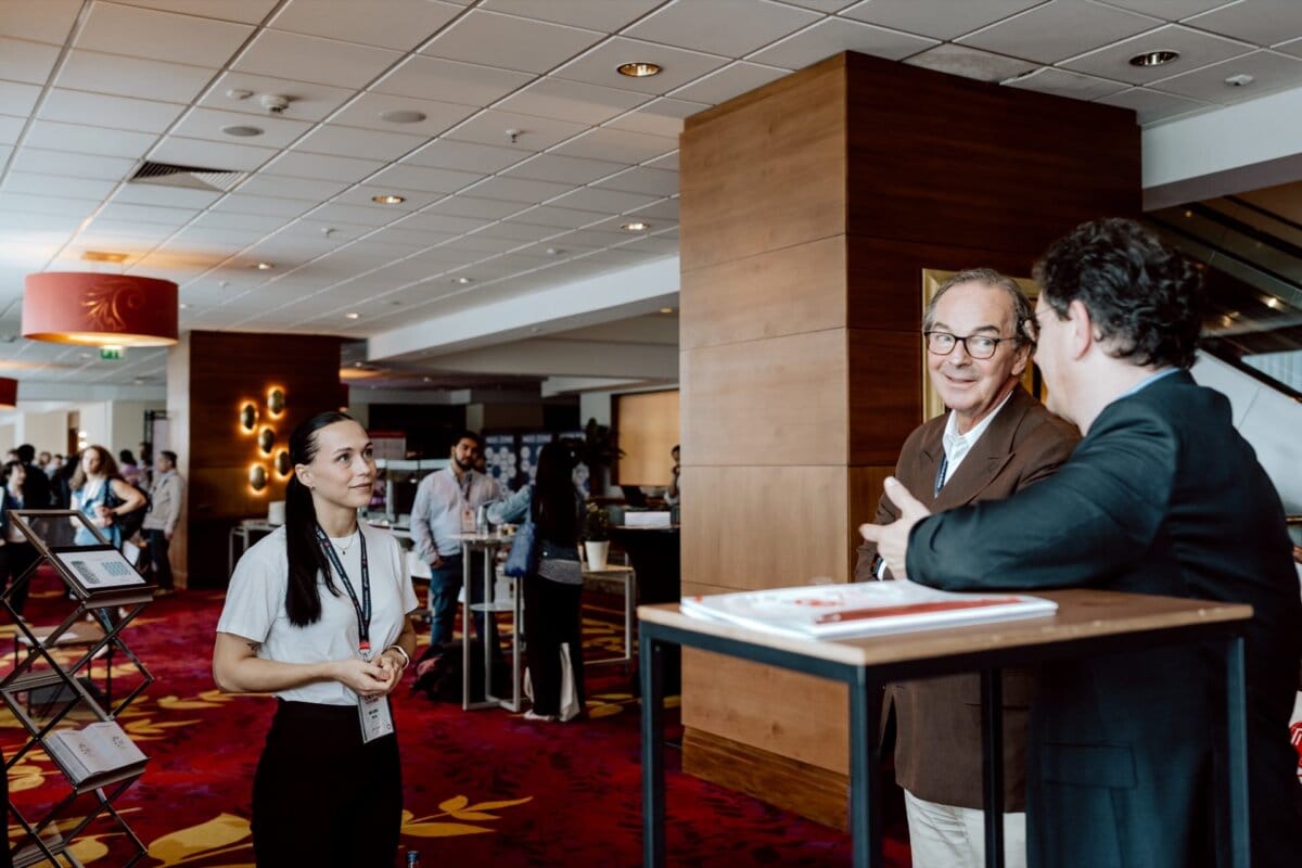 A woman is standing at an exhibition booth talking to two men dressed in business attire. They are in a conference room, surrounded by other attendees in the background. The environment is professional, focused on networking and discussion, capturing a photo-op of the conference.  