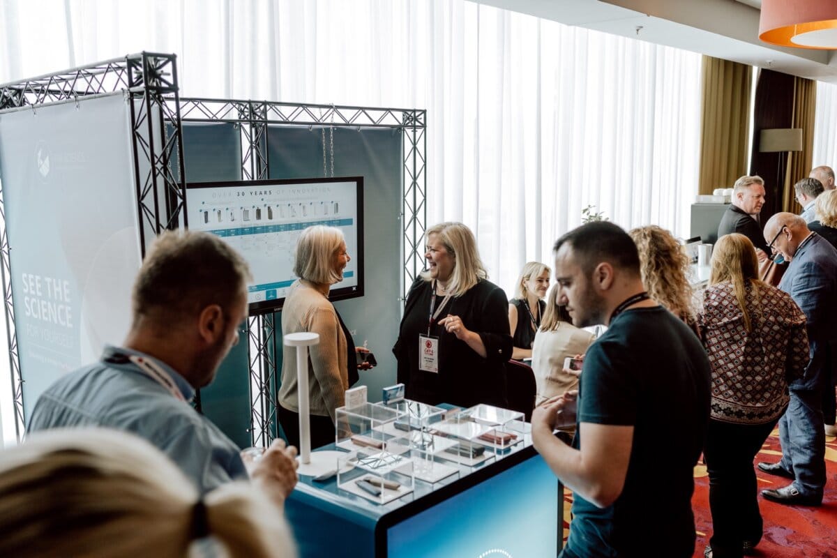 A group of people discuss at an exhibition booth during a conference. The booth is equipped with a screen and several transparent compartments displaying articles. Attendees, dressed in business attire, bask in the natural light streaming in through the large windows, making it an ideal place for photo coverage of the conference.  