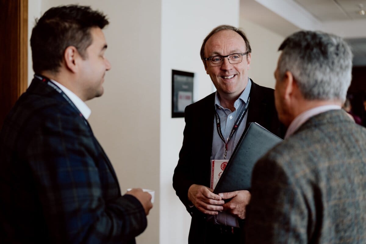 Three men attending a conference are engaged in conversation. The man in the middle, holding a briefcase, is smiling while the other two are listening intently. They are wearing business attire and give the impression that they are networking in a professional setting - the perfect moment for a photo essay on the conference.  