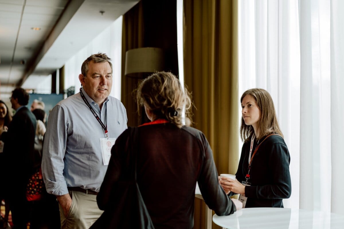 Three people are talking in a room with large windows. The man on the left is wearing a light blue shirt and a conference badge; opposite him are two women, one with her back to the camera, the other holding a cup, both also with badges - perfect for a photo essay of the event. 