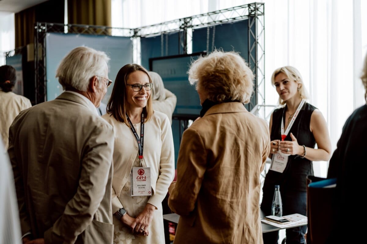 A group of people talking at a conference. A woman with glasses stands in the middle, smiling and wearing an attendee badge. Two other people make contact with her, while another woman stands with a drink in her hand. Exhibition booths are visible in the background, capturing the perfect moment for a reportage of the event.   