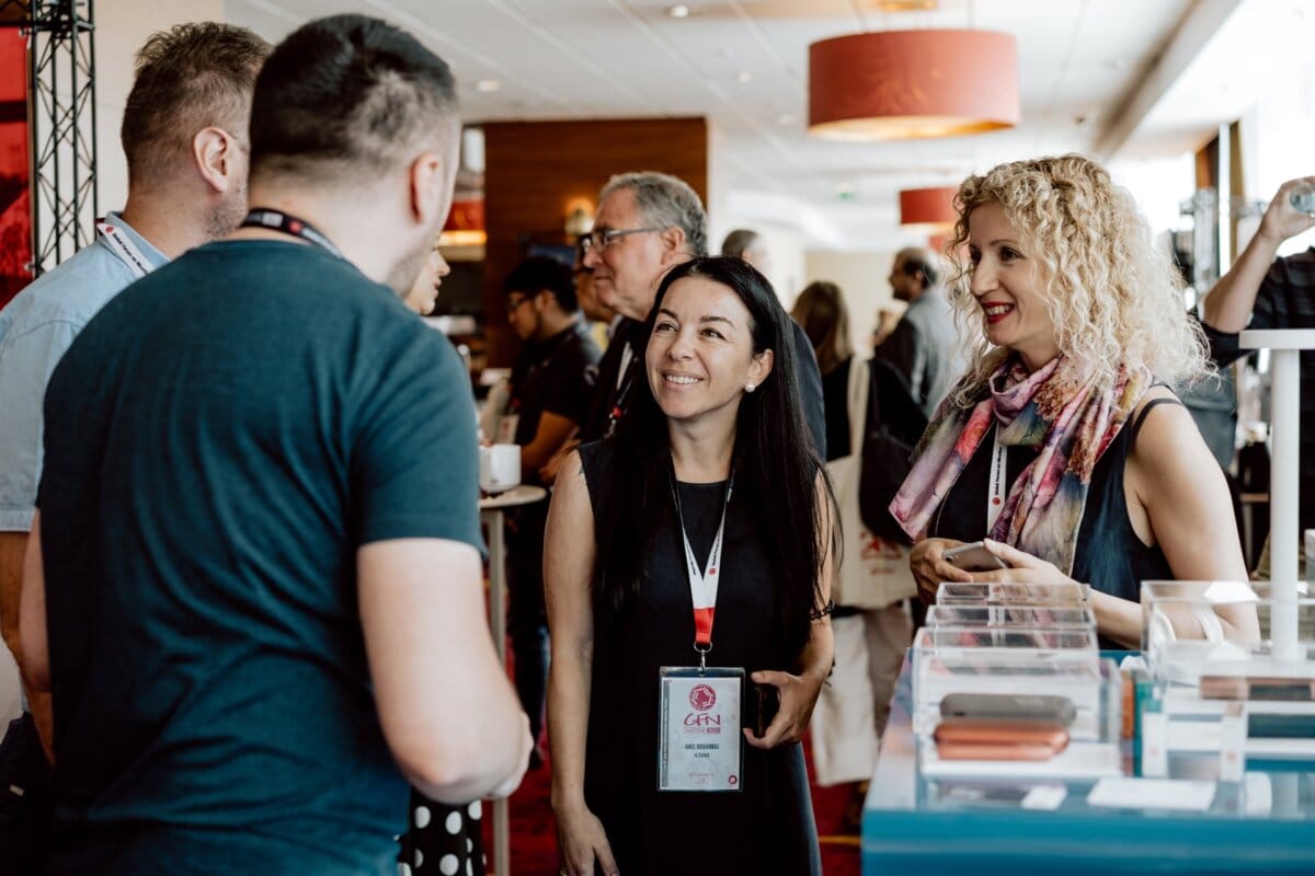 A group of people at a conference are having a conversation in a bright, modern room. Leading the way are two women and two men with conference badges. One woman with curly hair and a scarf smiles, while the other woman with long, dark hair listens intently - perfect moments for an event photographer capturing a photo essay of the event.  