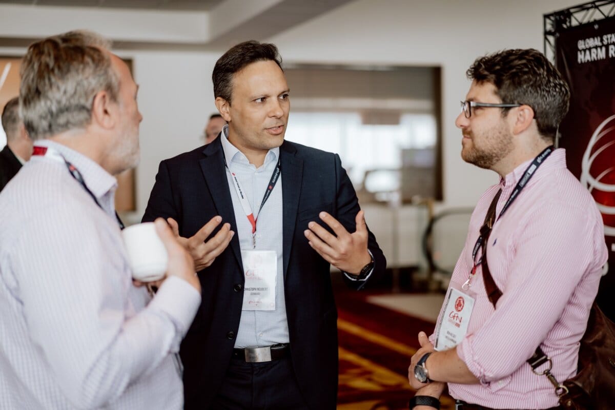 At a professional event, three men are having a conversation. The man in the middle, dressed in a suit, gestures with his hands. A white cup is held to his left; to his right, another wears glasses and a camera strap over his shoulder - presumably the event photographer documenting the occasion. All wear name badges.   