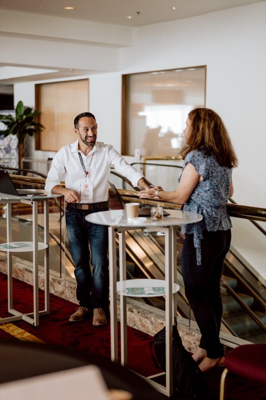 A man and a woman talk while standing at a high table, captured in a photo essay of the conference. The man, dressed in a white shirt and jeans, smiles warmly. The woman with long curly hair is wearing a patterned blouse and dark pants. Both appear to be in a modern, well-lit room.   