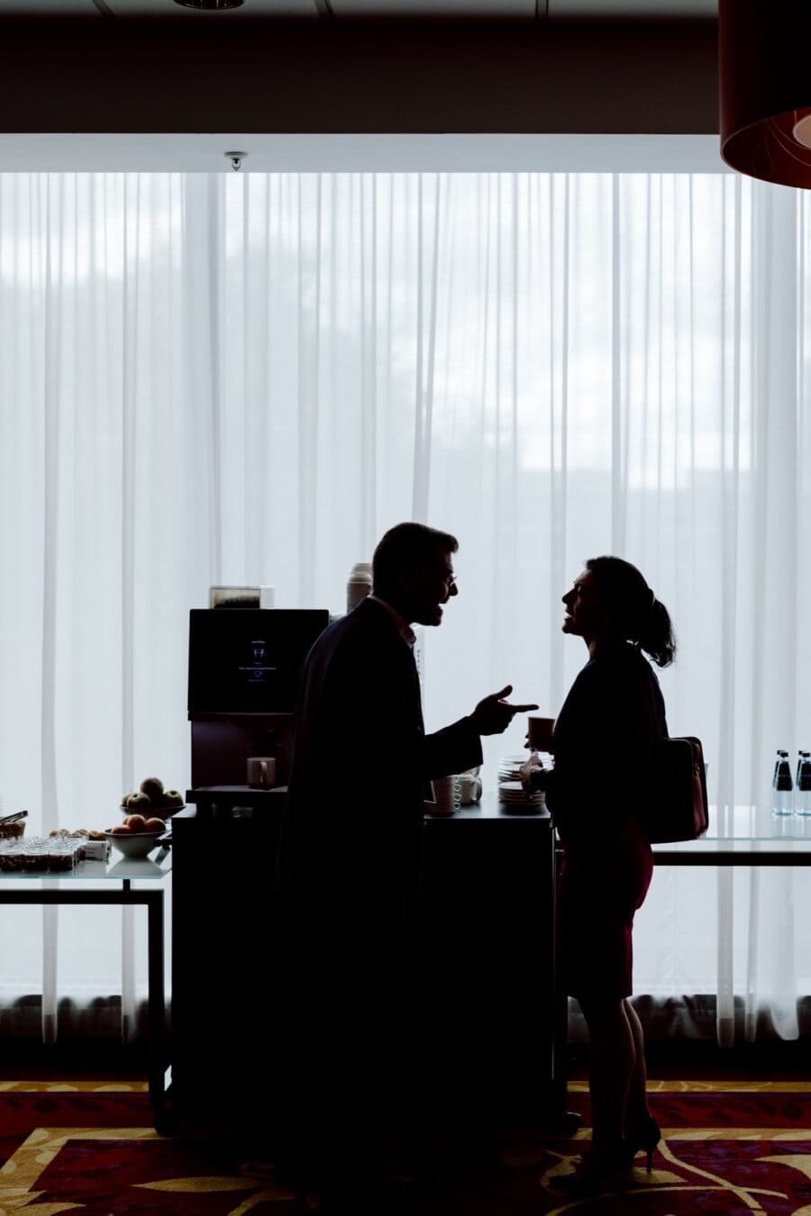 The silhouettes of a man and a woman stand facing each other, busy in conversation, against the backdrop of a bright window. Behind them is a coffee stand with a coffee maker, cups and an assortment of snacks. The room has a warm, softly lit atmosphere - the perfect place for an event photographer to capture every moment.  