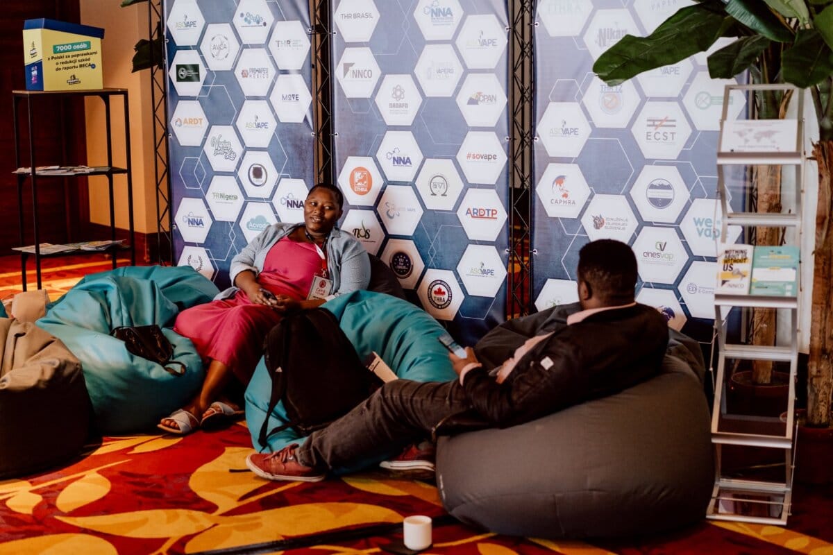 Two people are sitting across from each other on beanbags and are busy talking. In the background is a wall with various hexagonal logos and a tall plant on the right. A rack of brochures is also visible, capturing the essence of "event reportage" in this conference space.  