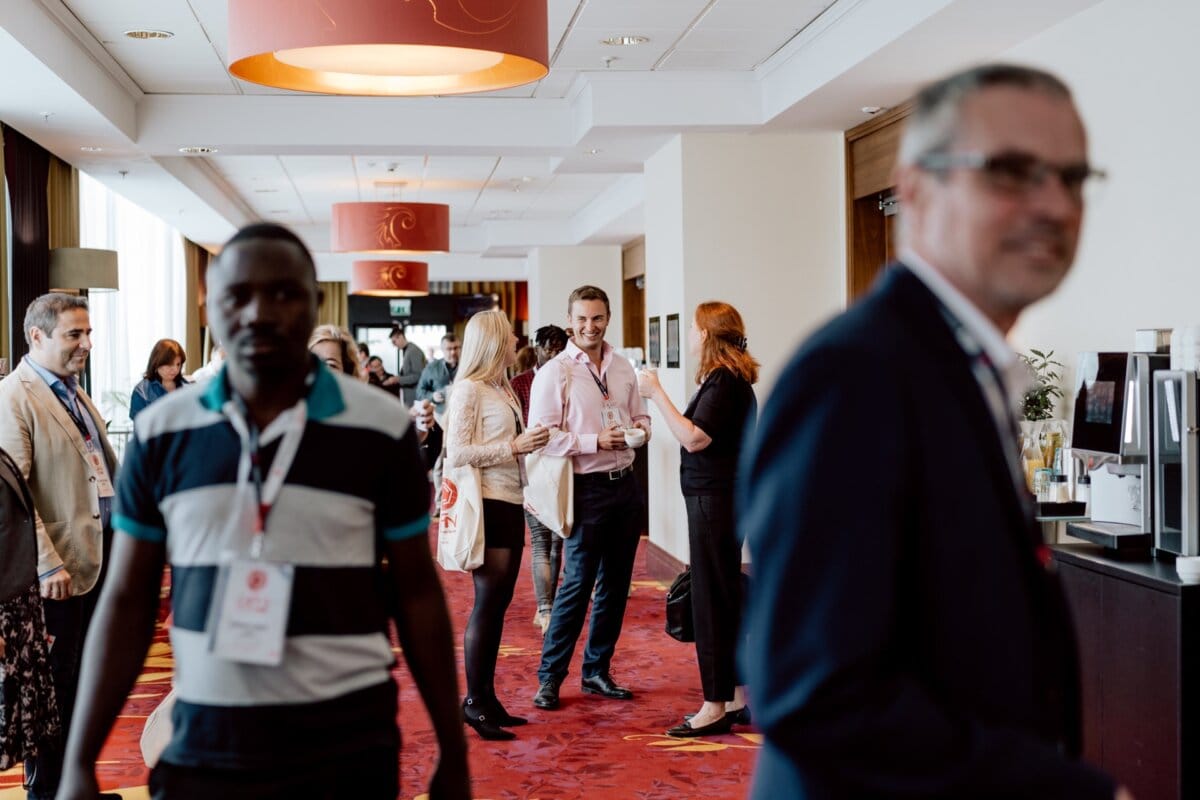 A group of people talking to each other in a brightly lit hallway during a conference, captured by a skilled event photographer. Some people are wearing name badges and holding cups of coffee. The atmosphere is relaxed and social, and the red patterned carpet and elegant lighting fixtures add to the charm.  