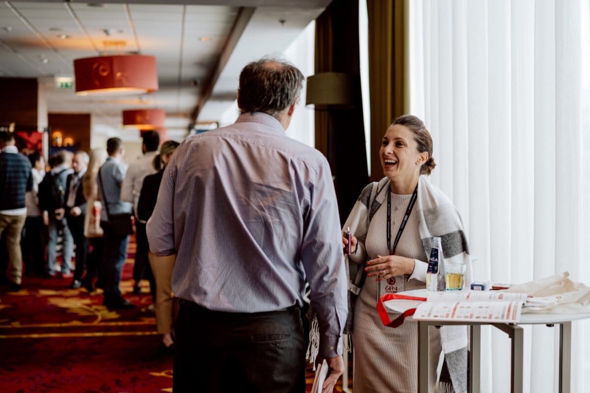 Two people are having a lively conversation in a conference room with large windows. In the background, numerous people form a line. The woman on the right is smiling and holding a lanyard and conference materials, the man seen from behind is listening attentively - the perfect moment for a photo essay of the event.  