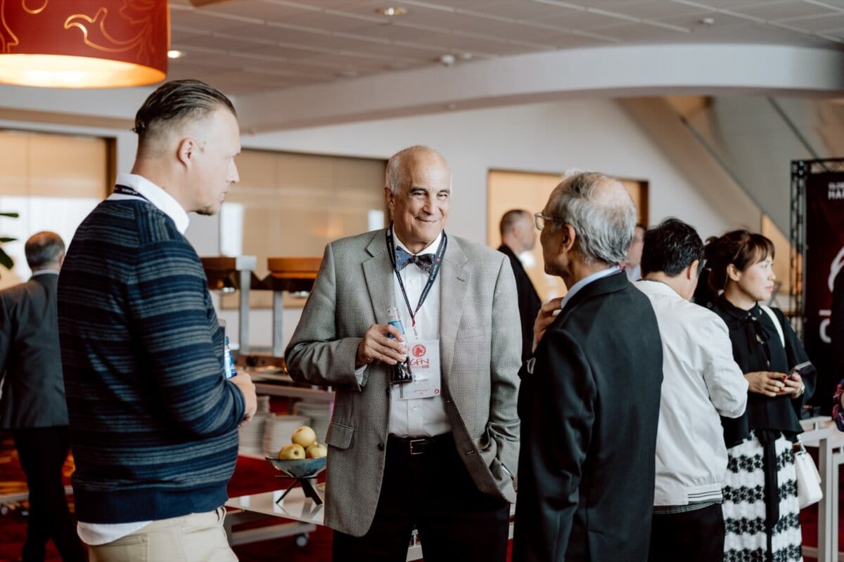 A group of people in a semi-formal setting, talking and holding drinks. Three men are talking in the foreground; one is holding a glass, the others seem attentive. Participants mingle at a buffet table in the background - perfectly captured in this photo essay of the event.  