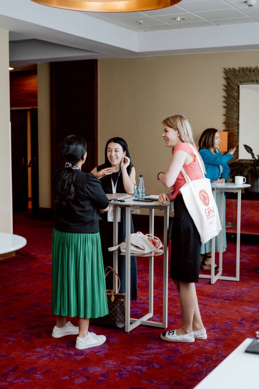 Three women are having a conversation at a high table set with conference badges and water bottles. One woman in a green skirt stands with her back to the camera, while two women face her smiling. In this photo essay from the conference, another person stands in the background at a similar table.  