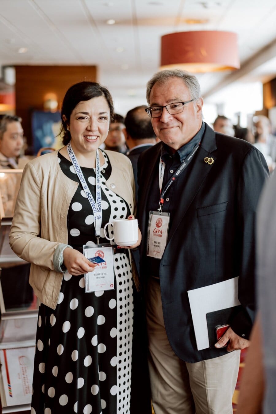 A woman in a polka dot dress holding a cup of coffee stands next to a man in a dark jacket, both with name badges around their necks. They are smiling and look as if they are attending a networking event or conference, which is perfectly captured in this photo essay of a conference among a crowd of business people. 