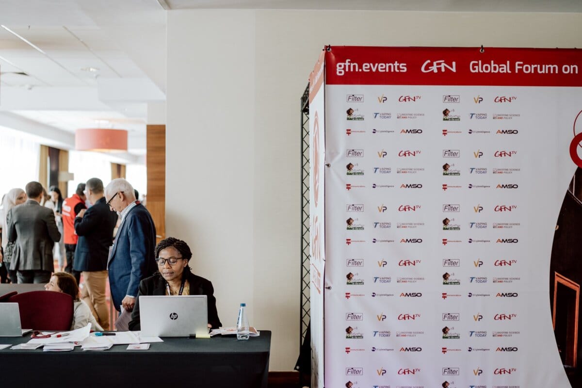 A person sits at a desk with a laptop in a conference room. To the right, a tall banner displays the logos of the Global Nicotine Forum and various sponsors. In the background, several people can be seen busy talking. The setting gives the impression of being professional and organized, ideal for photo coverage of the event.   