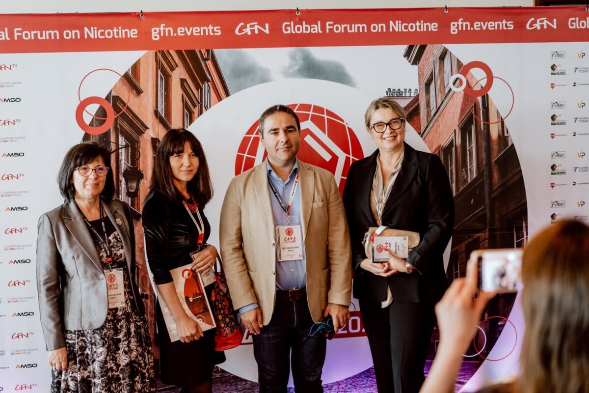 Four people are standing in front of the "Global Nicotine Forum" logo. They sit next to each other and smile at the camera. The person in the foreground takes a photo of them with a smartphone, capturing this snapshot of the conference. Conference badges can be seen on lanyards.   