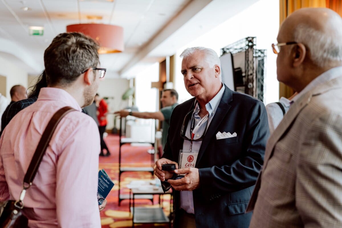 During a reportage of the event, three men can be seen carrying on a conversation in a professional atmosphere. One man, holding something in his hand, appears to be speaking to the others, who are listening intently. The background shows a spacious room with natural light and other people in the distance.  